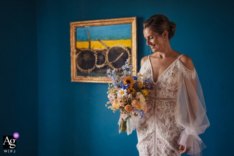 Castelnau des Fieumarcon, Lectoure, France — The bride gazes thoughtfully at her bouquet, standing indoors against richly painted dark blue walls, creating an elegant and serene wedding portrait.