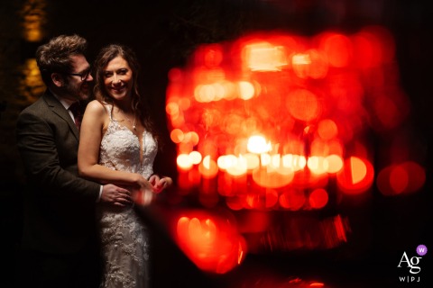 Dramatic Intimate Red Lit Wedding Portrait At Cripps Barn With Groom And Bride Smiling Cripps Barn — In a dark room lit with red gels, the groom stands behind the bride and leans in toward her smile, creating a dramatic and intimate wedding portrait at the barn venue.