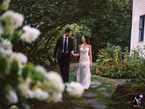 Warm Intimate Wedding Portrait - Coule Walking In Garden At The Barn At Walnut Hill Yarmouth Maine The Barn at Walnut Hill, Yarmouth — The couple walks hand in hand through the garden, looking at each other with the venue building visible on the right, creating a warm and intimate wedding portrait.