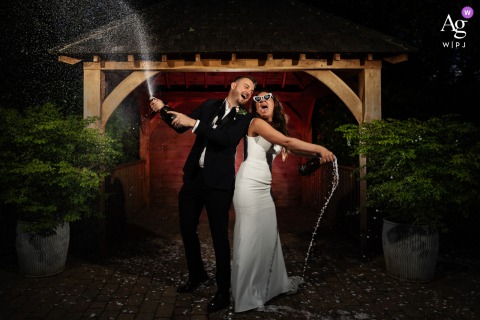Joyful Dynamic Champagne Spray Wedding Portrait At Tewinbury Garden With Timber Frame Structure Tewinbury — The couple smiles at each other while spraying champagne, lit by the photographer with a timber frame structure in the garden behind, creating a joyful and dynamic wedding portrait.