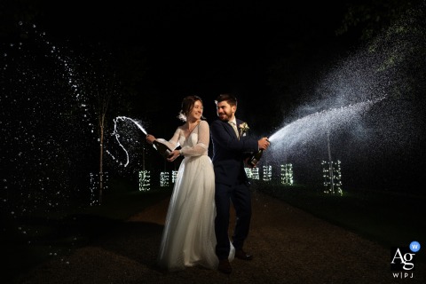 Celebratory Nighttime Champagne Spray Wedding Portrait At South Farm Dramatically Lit South Farm — The couple stands back to back, smiling as they spray champagne at night, dramatically lit by the photographer to create a lively and celebratory wedding portrait.