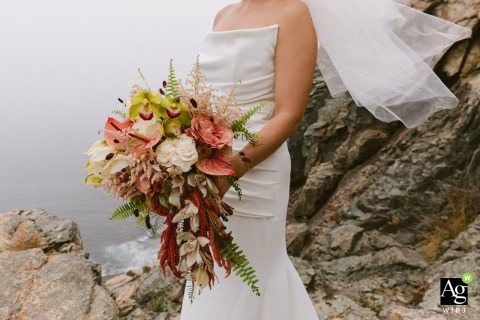 The bride holds her vibrant bouquet during a professional portrait session amidst the rugged coastal beauty and granite peaks of Acadia National Park in Maine.