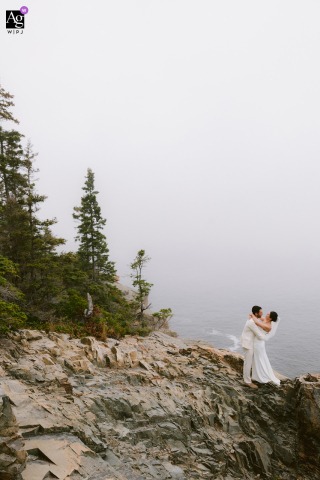 Acadia National Park — The bride and groom stand low in a tall, vertical minimalist frame on the cliffs, surrounded by fog, creating an ethereal and dramatic wedding portrait in the national park.