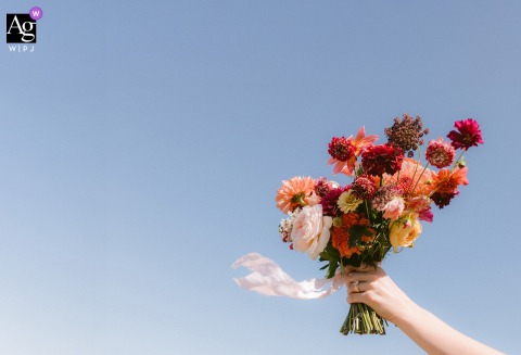 In South Portland, Maine, a minimalist detail shot captures the bride's hand and bouquet held high against a crisp blue sky, the flowers gently blowing in the wind on her wedding day.