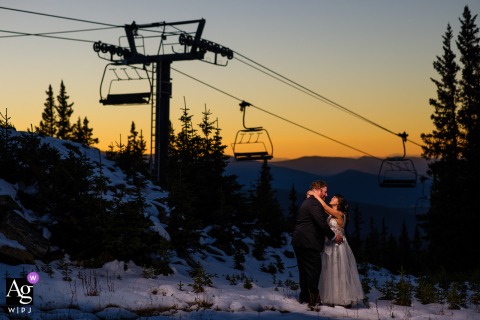 An evocative wedding portrait at Winter Park Resort shows the bride and groom positioned artistically beneath the strong geometric lines of a ski lift on a crisp, bright winter afternoon.