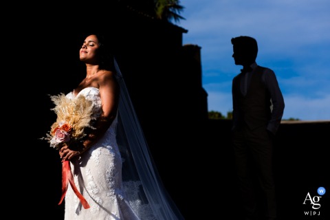 Domaine des Sources, Denicé, France — The bride basks in warm afternoon sunlight, taking a sun bath, while the groom appears in the background as a silhouetted, shadowed profile, creating an artistic wedding portrait at the reception venue.