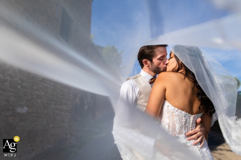 Domaine des Sources, Denicé, France — The married couple shares a kiss under the bride’s veil, romantically encircled by flowing tulle and bathed in warm afternoon sunlight at the reception venue.