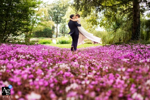 Pavillon d’Aymar, Saint-Romain-la-Motte, France — The groom lifts the bride playfully above a pink flowerbed at the reception venue, creating a joyful and whimsical wedding portrait set among colorful blooms.