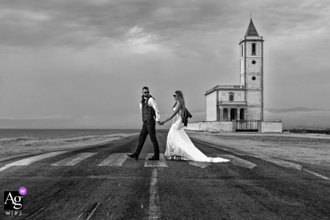 Cabo de Gata, Almería, Spain — The couple walks hand in hand across a crosswalk in a desolate setting, with a tall old structure in the background, captured in a black-and-white wedding portrait.
