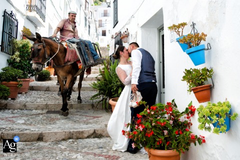 Frigiliana, Málaga, Spain — As the couple kisses in an urban village setting, a man rides past on a mule sitting side-saddle, adding a playful and authentic touch to their wedding portrait.