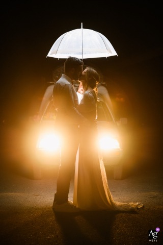 Toulouse, Haute-Garonne — The bride and groom stand under an illuminated umbrella in the reception venue car park, backlit by old car headlights, creating a dramatic and romantic wedding portrait at night.