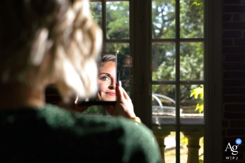 Domaine de Lalanne, Occitanie, France, a witness to the bride's preparation closely examines her makeup application mirrored in the reflection, highlighting the quiet anticipation before the ceremony.