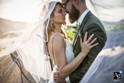 Clos de Florie, Savoie, France — During their sunset photo session, the bride and groom stand face to face, about to kiss beneath her sweeping veil that arcs across the entire frame in a romantic wedding portrait.