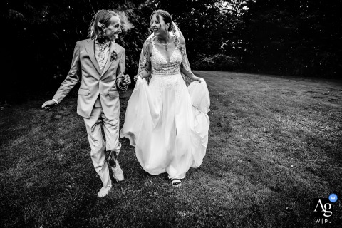 Gelderland, Netherlands — The bride and groom walk playfully outside on the grass in a black-and-white shot, unfazed as the rain begins to fall, capturing a carefree and joyful wedding portrait.
