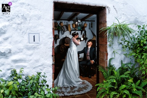 Frigiliana, Málaga, Spain — Captured from outside, the lit couple shares a playful indoor session as the bride pours from a bottle into the groom’s mouth, creating a fun and lively wedding portrait.