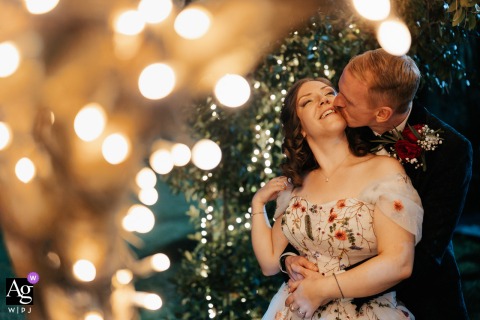 Villa Giulia, Vitorchiano (VT) — At night beneath hanging decorative lights, the groom kisses the bride’s neck from behind as she smiles, creating a romantic and warmly illuminated wedding portrait in the villa’s garden.