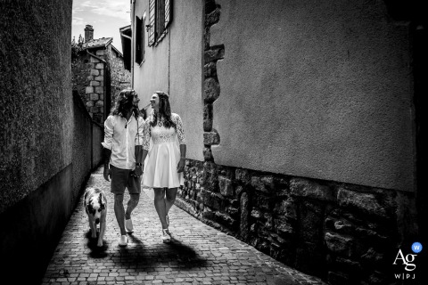 Brindas Rhône Newlyweds Walk Hand in Hand With Dog Sharing Kiss at Sunset Brindas, Rhône — The newlyweds walk hand in hand with their dog down an alleyway, sharing a kiss as the sun sets behind them, casting long shadows in this black-and-white, urban wedding portrait.