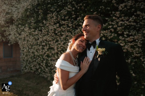San Giusto Abbey, Tuscania (VT) — The newlyweds, laughing in the sunlight, stand in front of a wall covered with lush plants and flowers as the bride gently rests her face on the groom’s chest in a relaxed wedding portrait.