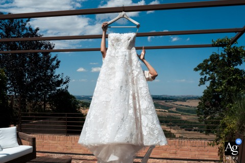 Macerata, Italy, a bride's white wedding dress hangs outside against a clear blue sky as a pair of hands gently reach to take it from the hanger.