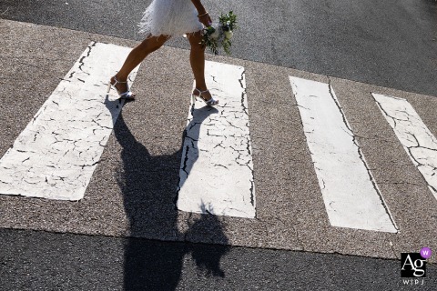 An artistic wedding day detail captured at the Mairie de Villefranque shows the bride's white gown and delicate shoes crossing the dark pavement and white striped crosswalk, highlighting the contrast in materials.
