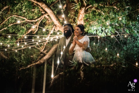 Recanto Coração de Jesus, Maceió - AL — The couple stands beneath a canopy of trees and strings of lights at night, their portrait beautifully reflecting the tree cover above for a romantic wedding image.
