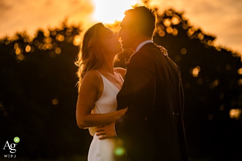 Sharing a romantic kiss at sunset, the newlyweds are silhouetted against the sky during a private portrait session held at the parents' house in the Hampshire, England countryside.