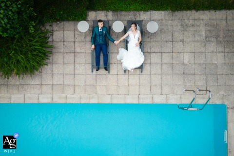 L'Héritage, Poliénas, France — The couple lies on lounge chairs at the edge of a bright blue square pool, captured in a creative drone portrait from above during their wedding session at the venue.