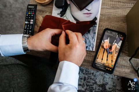 In Warwickshire, England, the groom's wedding morning preparation is captured in a detailed artistic close-up showing him referencing a YouTube tutorial on a mobile phone, a modern touch to the traditional rite.