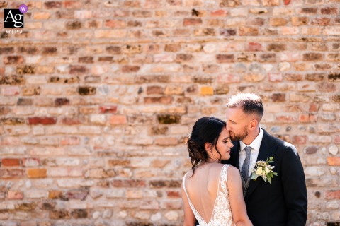 Udine, Italy — In a minimalist wedding portrait, the bride and groom are positioned low at the bottom of the frame as the groom kisses the bride's forehead, with a massive brick wall dramatically filling the background.