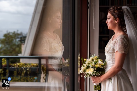 Trieste, Italy — The bride stands in profile on a balcony, near the window and holding her bouquet, captured in an elegant wedding portrait that highlights her serene expression and the soft natural light.