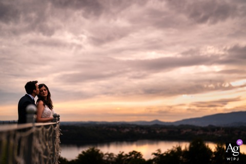 Villa Dolceacqua, Oggiono, Lake Como, Italy — The newlyweds pose in a minimalist sunset portrait, positioned low in the bottom left corner with a sweeping lake view and dramatic clouds filling the sky above them.