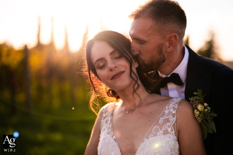 Udine, Italy — In a close-up sunset portrait, the groom stands behind the bride and kisses her cheek, with the golden glow illuminating the romantic outdoor wedding scene.