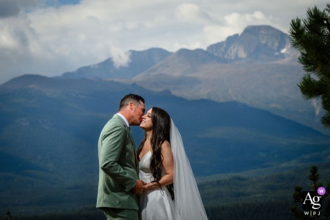 Rocky Mountain National Park — The groom kisses the bride on the cheek as they overlook a beautiful view, illuminated by the photographer, with dramatic white clouds adding depth to the scenic wedding portrait.