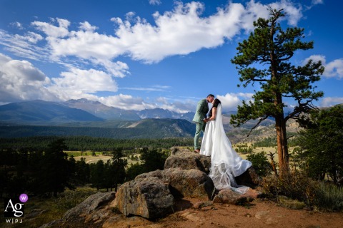 Rocky Mountain National Park, Estes Park, Colorado — The couple stands on a rock, kissing while illuminated by the photographer, with green trees, rolling hills, and a stunning mountain landscape framed by blue sky and white clouds above them.