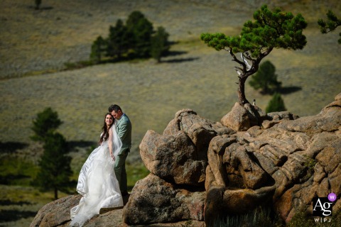 Rocky Mountain National Park, Estes Park, Colorado — The couple embraces on a windswept rock in a high-angle shot, with lush green trees and rolling hills providing a scenic backdrop to this artistic wedding portrait.