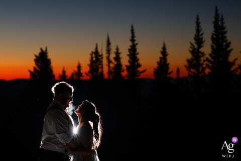 Lunch Rock at Winter Park Resort — The couple stands semi-silhouetted and rim lit by the photographer as the sun sets behind them, with the groom's breath visible in the cold mountain air for an artistic effect. 