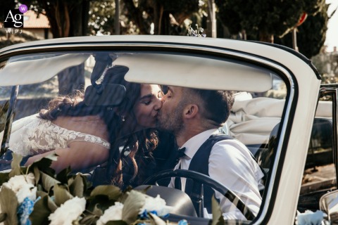 Through The Glass Of A Vintage Car At Santa Maria Del Castelvecchio Church In Savignano, The Newlyweds Share A Romantic Kiss Through the glass of a vintage car at Santa Maria del Castelvecchio Church in Savignano, Italy, the newlyweds share a romantic kiss, framed by classic automobile charm.