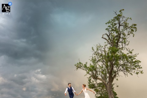The Couple Runs Under A Cloudy Sky At Their Dordogne Venue, With Dramatic Weather Emphasizing Mood And Open Landscape The couple moves quickly under a cloudy sky at their Dordogne reception venue, the dramatic weather emphasizing the mood and open landscape.