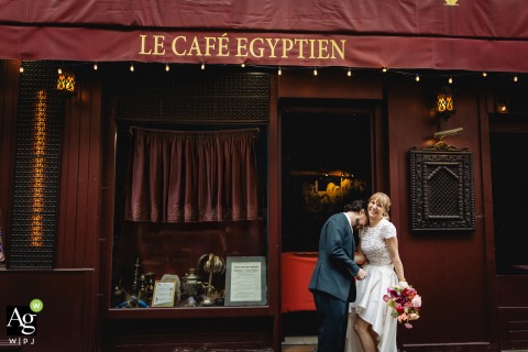 Bride and Groom Pose By Le Café Égyptien on Rue Mouffetard in Paris France On Rue Mouffetard in Paris, the bride and groom are photographed in front of the red façade of “Le Café Égyptien,” with the groom leaning against the bride’s shoulder while she holds her bouquet and smiles.