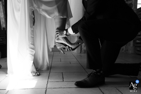 Inside Château Des Anglades In Hyères, France, The Best Man Assists The Bride With Her Shoes In A Tender Wedding Detail Inside Château des Anglades in Hyères, France, the best man helps the bride put on her shoes, highlighting a tender and supportive wedding day detail.