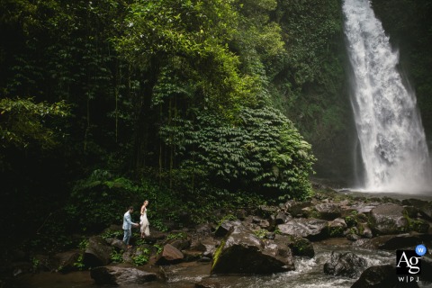 In The Jungle Of Bali, Indonesia, The Couple Walks Toward A Giant Waterfall With Lush Greenery And Rushing Water In the jungle of Bali, Indonesia, the couple walks toward a giant waterfall, with lush greenery and rushing water creating a breathtaking natural backdrop.