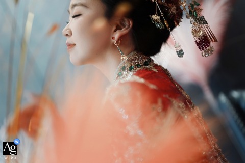 In Her Qidong Home, The Bride Glows In A Red Chinese Wedding Gown With Ornate Hairpieces And Decorative Feathers In her home in Qidong, China, the bride glows in a red traditional Chinese wedding gown and ornate hairpieces, her profile softly outlined against decorative feathers.