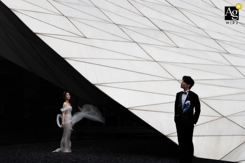 At The Entrance Of A Hotel In Nanping, Fujian, The Couple Stands Beneath Symmetrical Structure For A Striking Portrait At the entrance of a hotel in Nanping, Fujian, the bride and groom stand beneath the building’s symmetrical structure, creating a visually striking wedding portrait.
