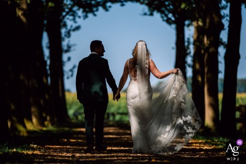 On The Grounds Of Kasteel Dussen In Zuid Holland, Netherlands, The Couple Walks Hand In Hand Through Beautiful Natural Sunlight On the picturesque grounds of Kasteel Dussen in Zuid Holland, Netherlands, the bride and groom walk hand in hand, illuminated by beautiful natural sunlight.