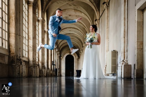 The Groom Leaps Energetically In A Light Blue Suit In The Vaulted Corridor At Abbaye De Prémontrés, Partner Smiling At Abbaye de Prémontrés, a groom in a light blue suit leaps energetically in a vaulted corridor, while his partner, holding her bouquet, looks on with a smile.