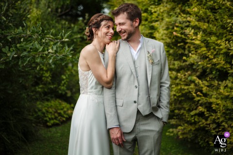 In A Metz Garden, The Bride With Braided Hair Smiles Tenderly At Her Groom In A Light Gray Suit In a lush garden at the reception venue in Metz, France, the bride with braided hair smiles tenderly at her groom in a light gray suit as he laughs joyfully.