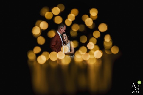 Golden Bokeh Lights in an Intimate Forehead Kiss Portrait at Domaine de Raville in Moselle Bathed in a halo of blurred, bokeh golden lights at Domaine de Raville in Moselle, a groom gently kisses his bride’s forehead as they share an intimate, tender exchange in the darkness.