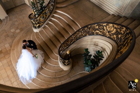 The Bride And Groom Share A Peaceful Kiss At The Base Of A Monumental Staircase In Nancy, France In Nancy, France, the bride and groom share a peaceful moment at the base of a monumental staircase, the sweeping architecture lending elegance to their portrait.