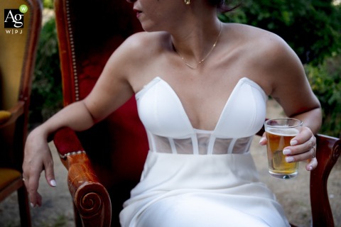 Seated Bride has a Beer to Celebrate and Embrace Joyful Atmosphere in Largentiere France Enjoying a laid-back celebration in Largentiere, France, the bride raises a beer to the festive spirit and the relaxed, joyful atmosphere of the day.