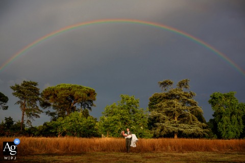 The Groom Carries The Bride Beneath A Rainbow At Château Destinée, France, Creating A Wonder-Filled Wedding Portrait In a picturesque field below a rainbow and stormy skies at Château Destinée, France, the groom carries the bride, the vibrant scenery adding a sense of wonder to their portrait.